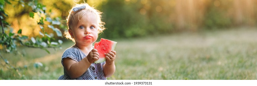 Cute Caucasian baby girl eating ripe red watermelon in park. Funny child kid with fresh fruit outdoors. Supplementary healthy finger food for toddler kids. Summer fun. Web banner header.