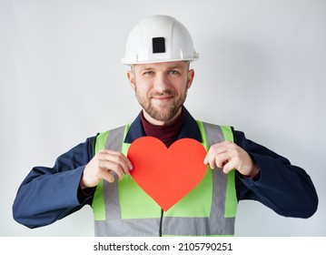 Cute Caucasian Bearded Construction Supervisor, Contractor Or Engineer Standing By The Wall Holding Red Heart Shape. Worker With Paper Heart. Romance, Relationships Concept. Valentine's Day Symbol