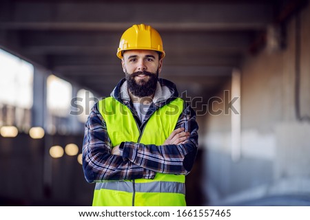 Cute Caucasian bearded construction worker with safety helmet on head in vest standing with arms crossed at construction site and looking at camera.