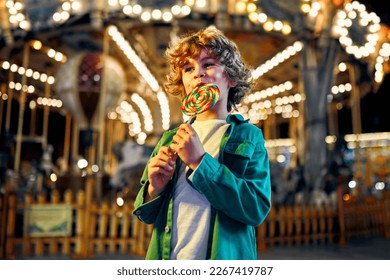 A cute caucasian boy with blonde curly hair eating a colorful lollipop standing against the background of a carousel with horses in the evening at an amusement park or circus.