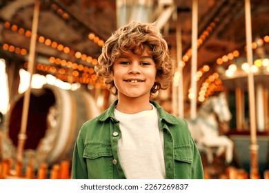 A cute Caucasian boy, a child with curly hair, came to the amusement park to have fun, play and ride a merry-go-round. A child standing in front of a merry-go-round with horses. - Powered by Shutterstock