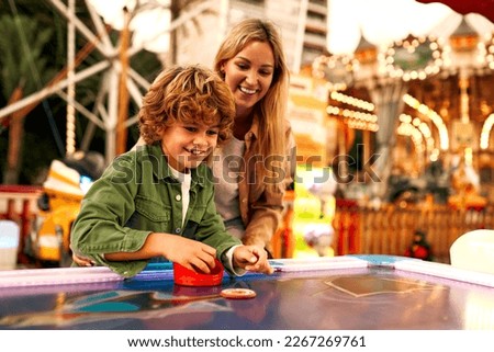 A cute Caucasian boy with curly hair with his mother playing with a puck in an air hockey in an amusement park and carousel on a weekend.
