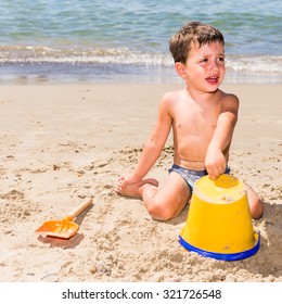 Cute Caucasian Child Crying While Playing On The Beach With Pail And Shovel In A Sunny Day With Sea Background