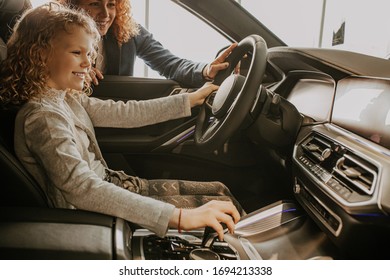 Cute Caucasian Child Girl Sitting At The Wheel Of A New Buisness Class Car In Dealership. Girl's Parents Going To Buy New Car, Make Purchase