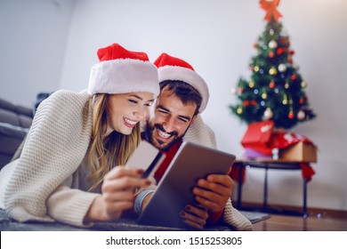 Cute Caucasian couple lying on stomach on the floor with santa hats on heads and using tablet for online shopping. Woman holding credit card. Living room interior. In background is christmas tree. - Powered by Shutterstock