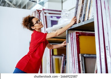 Cute Caucasian Female Employee With Curly Hair And With Eyeglasses Reaching For Silk Screen Plates. Printing Shop Interior.