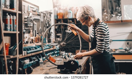 Cute Caucasian female worker holding and repairing bicycle wheel while standing in bicycle workshop. - Powered by Shutterstock