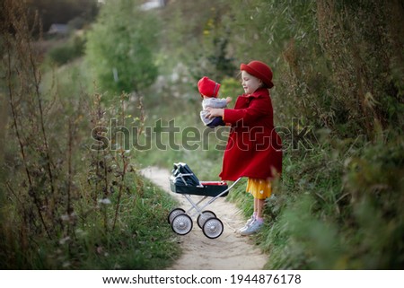 Cute caucasian girl in a red coat and hat with baby doll and doll stroller in the park, a child like mom, girl playing with doll, taking care of doll, role play and motherhood concept, little lady