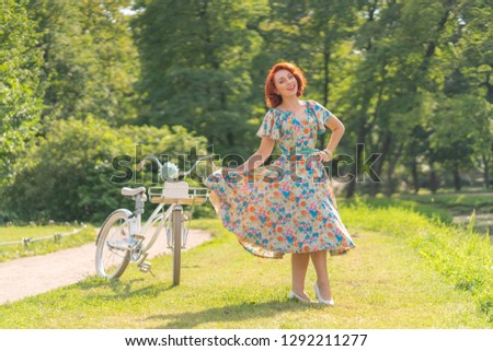 cute Caucasian girl with red hair in a retro dress enjoying life with a white vintage Bicycle in sporting fashionable walk in a city Park at summer day