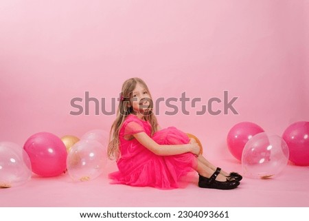 Cute Caucasian girl of seven years old in a bright pink barbicore style dress sits on a pink background with festive water balloons for her birthday