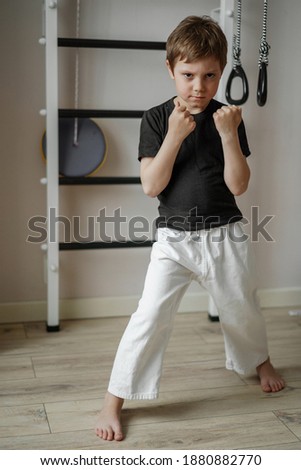 cute caucasian karate boy standing in stance in white kimono pants and black tee shirt at home near wall bars. Individual martial art sport for kids.