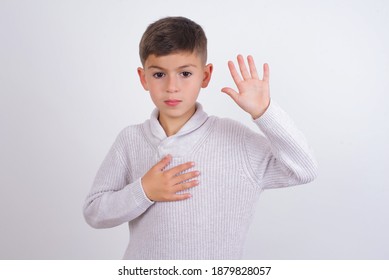 Cute Caucasian Kid Boy Wearing Knitted Sweater Against White Wall Swearing With Hand On Chest And Open Palm, Making A Loyalty Promise Oath