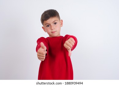 Cute Caucasian Kid Boy Wearing Red Knitted Sweater Against White Wall Feeling Unsure Making Good Bad Sign. Displeased And Unimpressed.