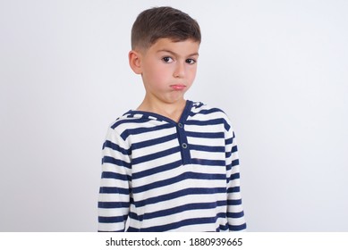 Cute Caucasian kid boy wearing stripped knitted sweater against white wall with snobbish expression curving lips and raising eyebrows, looking with doubtful and skeptical expression, suspect and doubt