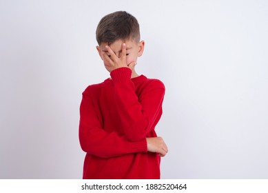 Cute Caucasian Kid Boy Wearing Knitted Sweater Against White Wall Making Facepalm Gesture While Smiling Amazed With Stupid Situation.