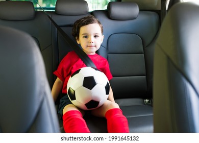 Cute Caucasian Kid Boy Wearing A Red Uniform And Holding A Soccer Ball In The Car. Parents Driving His Son To A Football Practice  