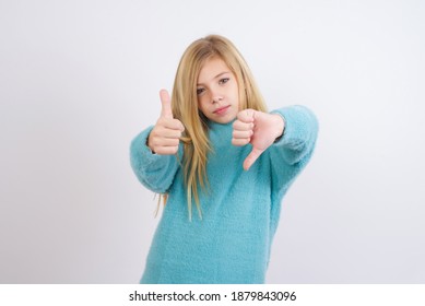 Cute Caucasian Kid Girl Wearing Blue Knitted Sweater Against White Wall Feeling Unsure Making Good Bad Sign. Displeased And Unimpressed.