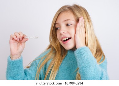 Cute Caucasian kid girl wearing blue knitted sweater against white wall holding an invisible braces aligner. Dental healthcare concept. 