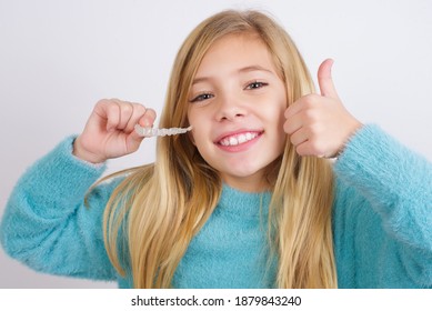 Cute Caucasian Kid Girl Wearing Blue Knitted Sweater Against White Wall Holding An Invisible Braces Aligner And Making Ok Sign With Hand. Dental Healthcare Concept. 