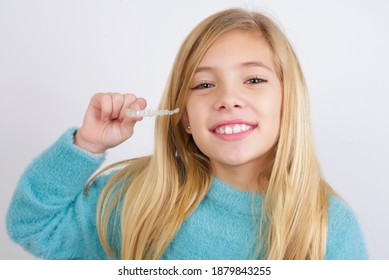 Cute Caucasian Kid Girl Wearing Blue Knitted Sweater Against White Wall Holding An Invisible Braces Aligner. Dental Healthcare Concept. 