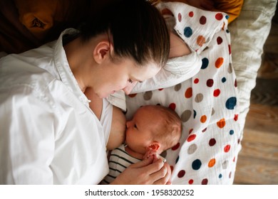 Cute caucasian mom and newborn baby, mother breastfeeds the baby lying on the bed in a light white bedroom with a muslin diaper. Top view, toning and lifestyle in a real interior