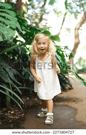 Cute caucasian pretty blonde two year old girl, toddler, kid among big green exotic leaves laughing looking at camera. Natural beauty,perfect skin, happy childhood concept.Full body shot.
