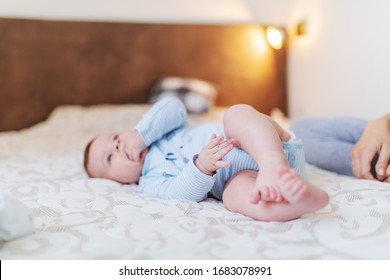 Cute Caucasian Six Months Old Baby Boy Dressed In Bodysuit Lying On Bed In Bedroom And Sucking Fingers. Next To Him Is Mother.