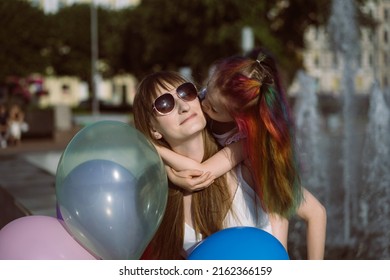 Cute Caucasian Smiling Girl With Colorful Dyed Hair Kissing Mom On Cheeck Sitting By Fountain. Image With Selective Focus. 