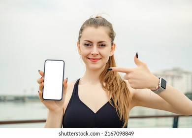 Cute Caucasian Woman Wearing Sportive Clothes On City Park, Outdoors Holding A Phone And Pointing Finger To Phone With Smiley Face. She Is Looking To Cell Phone Screen.