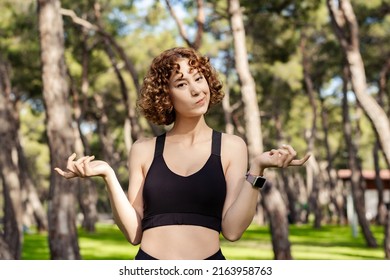 Cute Caucasian Woman Wearing Sports Bra Standing On City Park, Outdoor Clueless And Confused With Open Arms, Shrugging Shoulders, No Idea And Doubtful Face.