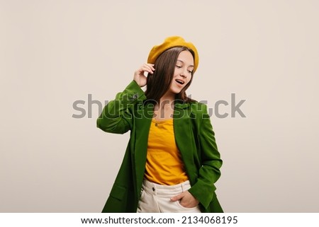 Cute caucasian young girl with parted mouth looks to side on white background. Brunette touches her hair and wears yellow beret, blouse and green jacket. Lifestyle and fashion concept.