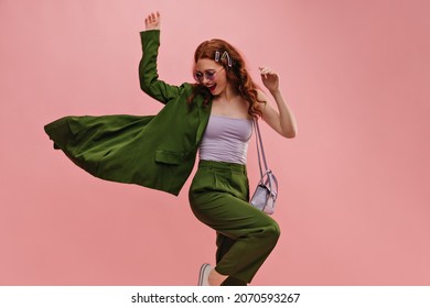 Cute caucasian young girl vigorously posing waving jacket to side in photo studio. Red curly-haired lady is dressed in lilac top, green suit and bag. Leisure concept of youth