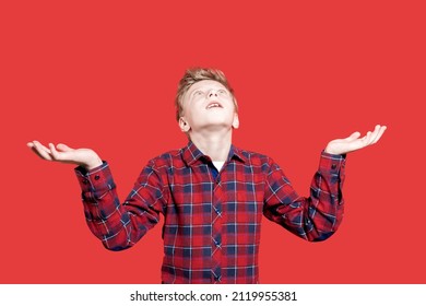 Cute Caucasian Young Teen Smiling And Looking Up, Isolated On A Red Background. Child Is Fun. Catches Snow. Portrait Of A Teenage Boy In A Red Shirt, Raising His Hands And Trying To Catch Something.