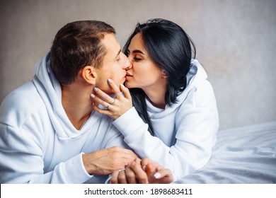Cute, charming, attractive young Europeans man and woman lie relaxed on the bed. Looking out the window, hug, stroke each other. Foreheads, faces together, kiss. Tenderness at home.