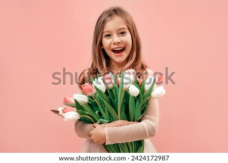 Cute charming little girl in a pink dress holds a bouquet of tulips flowers isolated on a pink background.