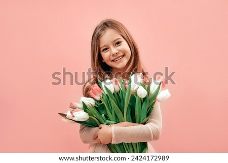 Cute charming little girl in a pink dress holds a bouquet of tulips flowers isolated on a pink background.