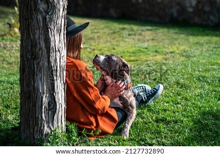 A cute and cheerful Caucasian woman in a hat is sitting in the park next to her dog of the American Stafford Terrier breed