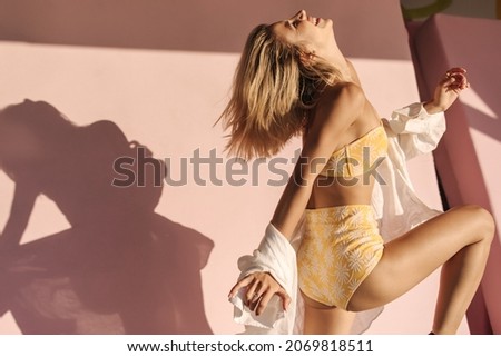 Cute cheerful caucasian young woman tilting her head back laughing closing her eyes on pink background. Blonde with bob haircut in swimsuit and shirt presses her leg to knee.
