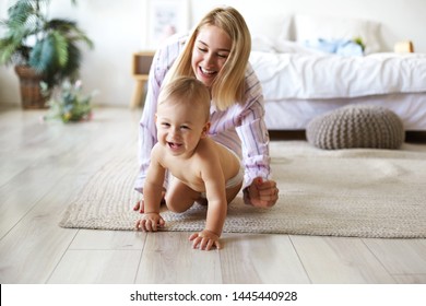Cute cheerful European infant in diaper having joyful facial expression, laughing while crawling on floor from his smiling mother who is chasing him. Blonde young woman playing with son indoors