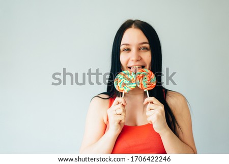 A cute cheerful girl in a bright orange t-shirt is having fun and nibbling colorful lollipops on sticks