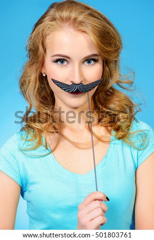 Cute cheerful girl holding fake mustache on a stick and smiling. Studio shot. 
