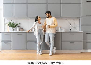 Cute cheerful loving young indian man and woman wearing homewear standing by table, drinking coffee together at kicthen in the morning, have conversation, smiling, full length, copy space - Powered by Shutterstock