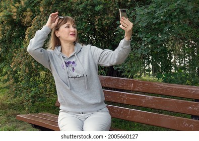 Cute Cheerful Middle Aged Blonde Sitting On A Park Bench Looking At The Phone, Outdoor Portrait Of A Mature 45 Year Old Woman Taking A Selfie In The Park.