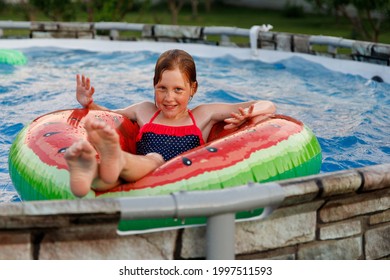 A Cute Cheerful Red-haired Girl 10 Years Old Swims In A Frame Pool On A Swimming Ring In Summer In Sunny Weather In The Backyard Of A Private House.