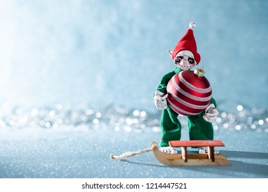 Cute Cheerful Santas Helper Elf Loading Christmas Bauble Onto Santas Sleigh. North Pole Christmas Scene. Santas Workshop. Elf At Work. 