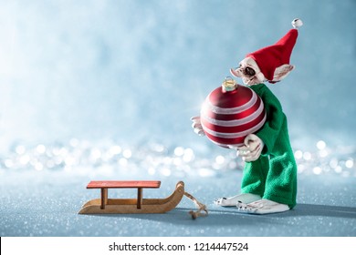 Cute Cheerful Santas Helper Elf Loading Christmas Bauble Onto Santas Sleigh. North Pole Christmas Scene. Santas Workshop. Elf At Work. 