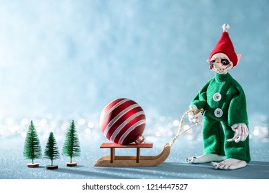 Cute Cheerful Santas Helper Elf Pulling Santas Sleigh With Christmas Bauble.North Pole Christmas Scene. Santas Workshop. Elf At Work. 