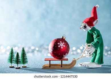 Cute Cheerful Santas Helper Elf Pulling Santas Sleigh With Christmas Bauble.North Pole Christmas Scene. Santas Workshop. Elf At Work. 