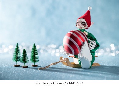 Cute Cheerful Santas Helper Elf Sitting On Santas Sleigh With Christmas Bauble. North Pole Christmas Scene. Santas Workshop. Elf At Work.