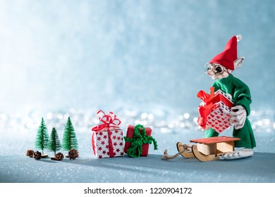 Cute Cheerful Santas Helper Elf Loading Christmas Gifts Onto Santas Sleigh. North Pole Christmas Scene. Santas Workshop. Elf At Work. 